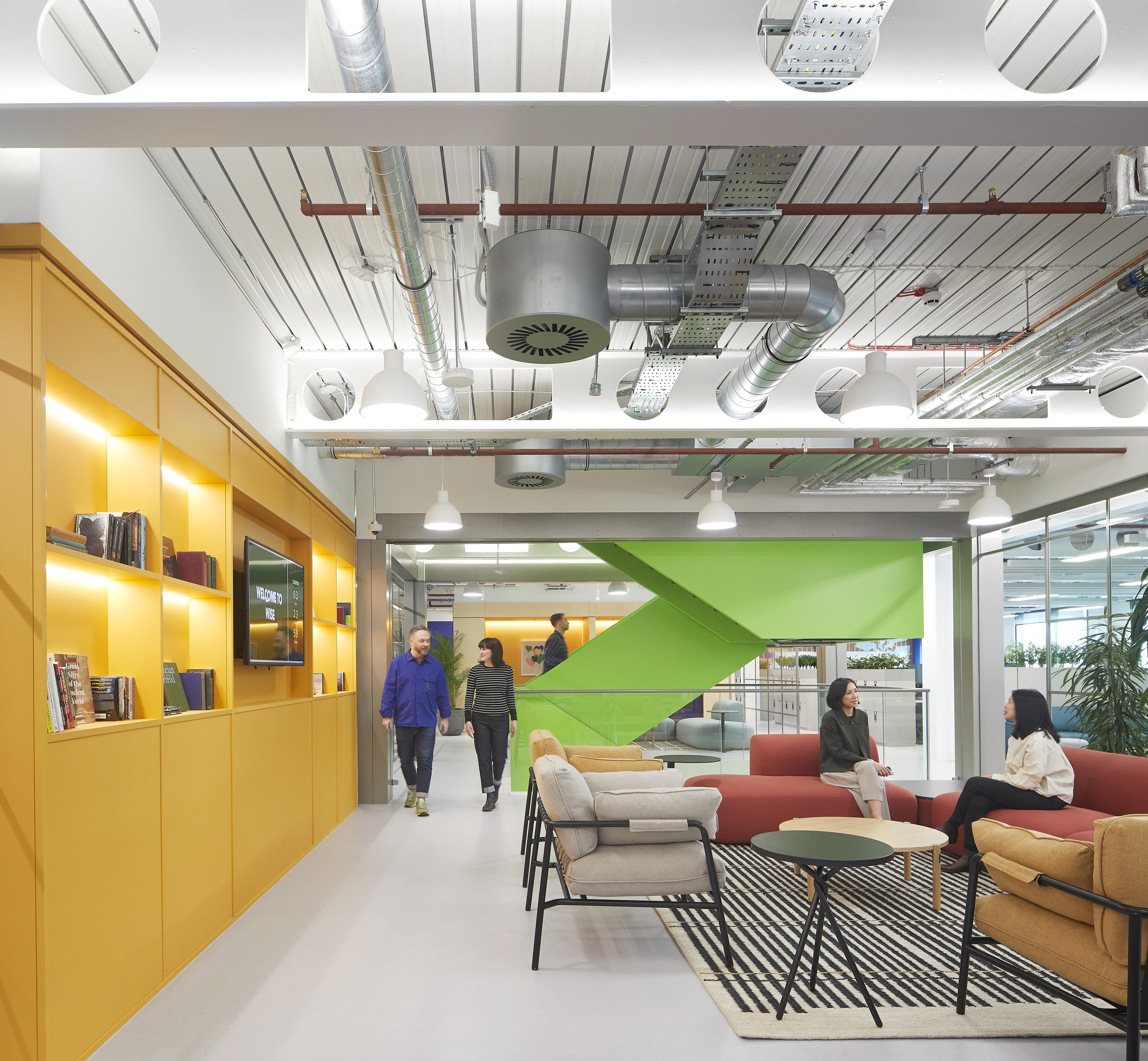 Open plan office fit out with a modern breakout area featuring colourful sofas and armchairs, built-in shelving, exposed ceiling services and suspended lighting, with staff using the space for informal meetings within a contemporary office design.