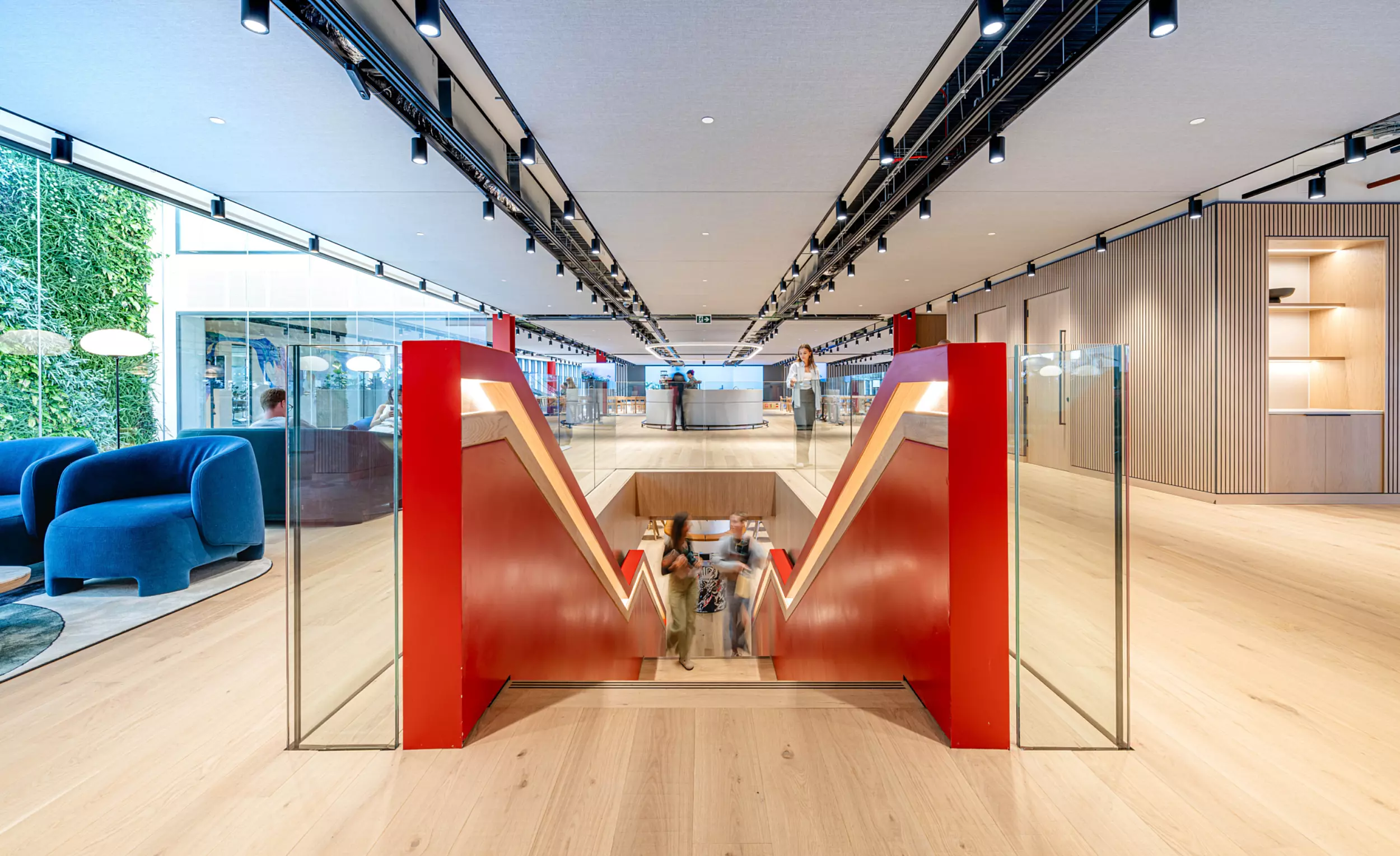 A modern office space with a central red staircase leading downwards. The area is well-lit with ceiling lights and features wooden flooring. On the left there are blue armchairs and a green wall with plants. On the right there is a wooden panelled wall with built-in shelves. People can be seen using the staircase and walking around the space.