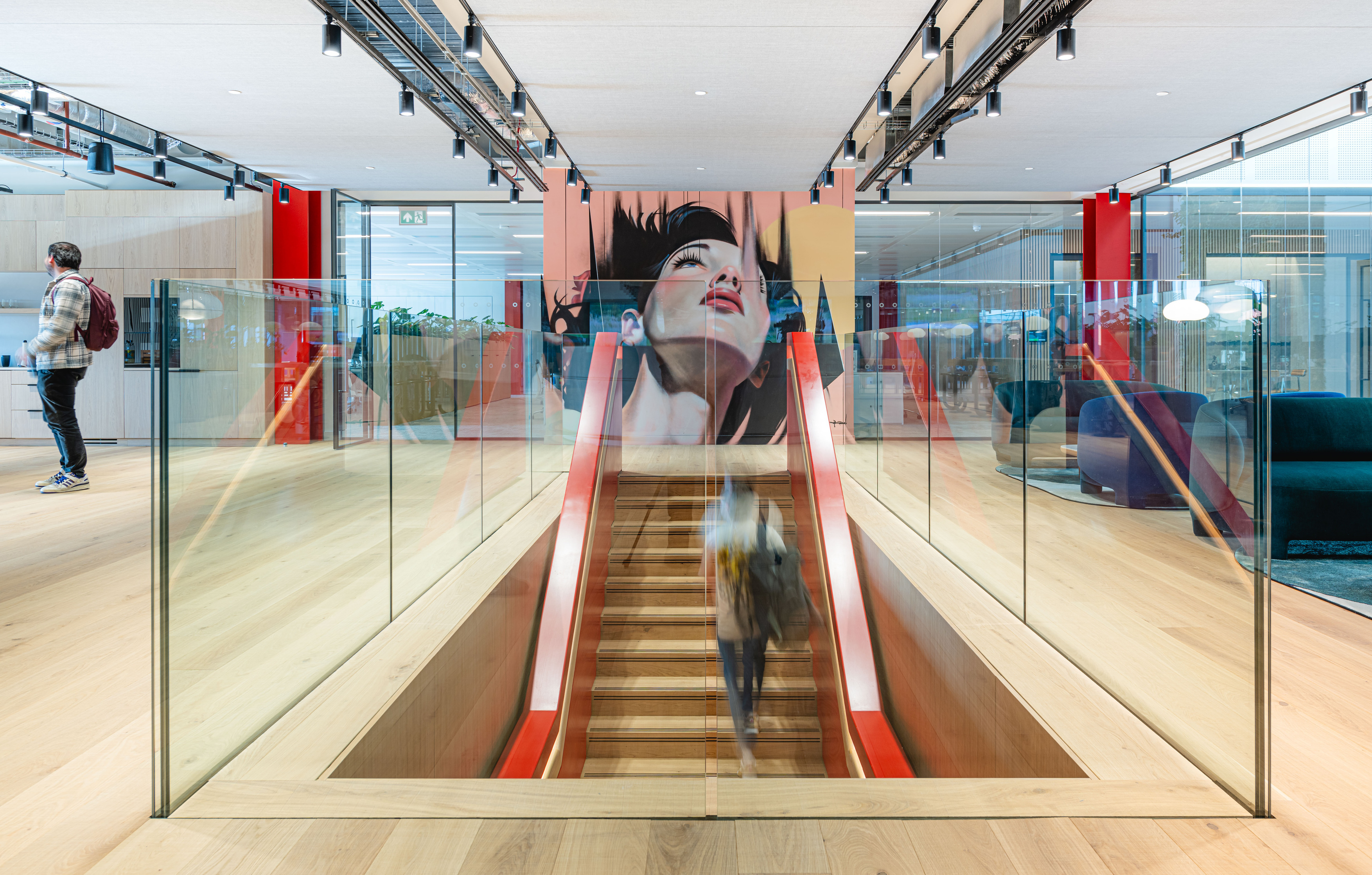 The image shows a modern office fit out and office design. It features an open-plan layout with wooden flooring and glass railings. A staircase with red handrails leads to a lower level, bordered by glass panels. At the top of the stairs is a large mural of a woman's face, adding an artistic element to the space. The ceiling has track lighting and there are several seating areas with contemporary furniture in the background. The overall design highlights openness, light and modern aesthetics.