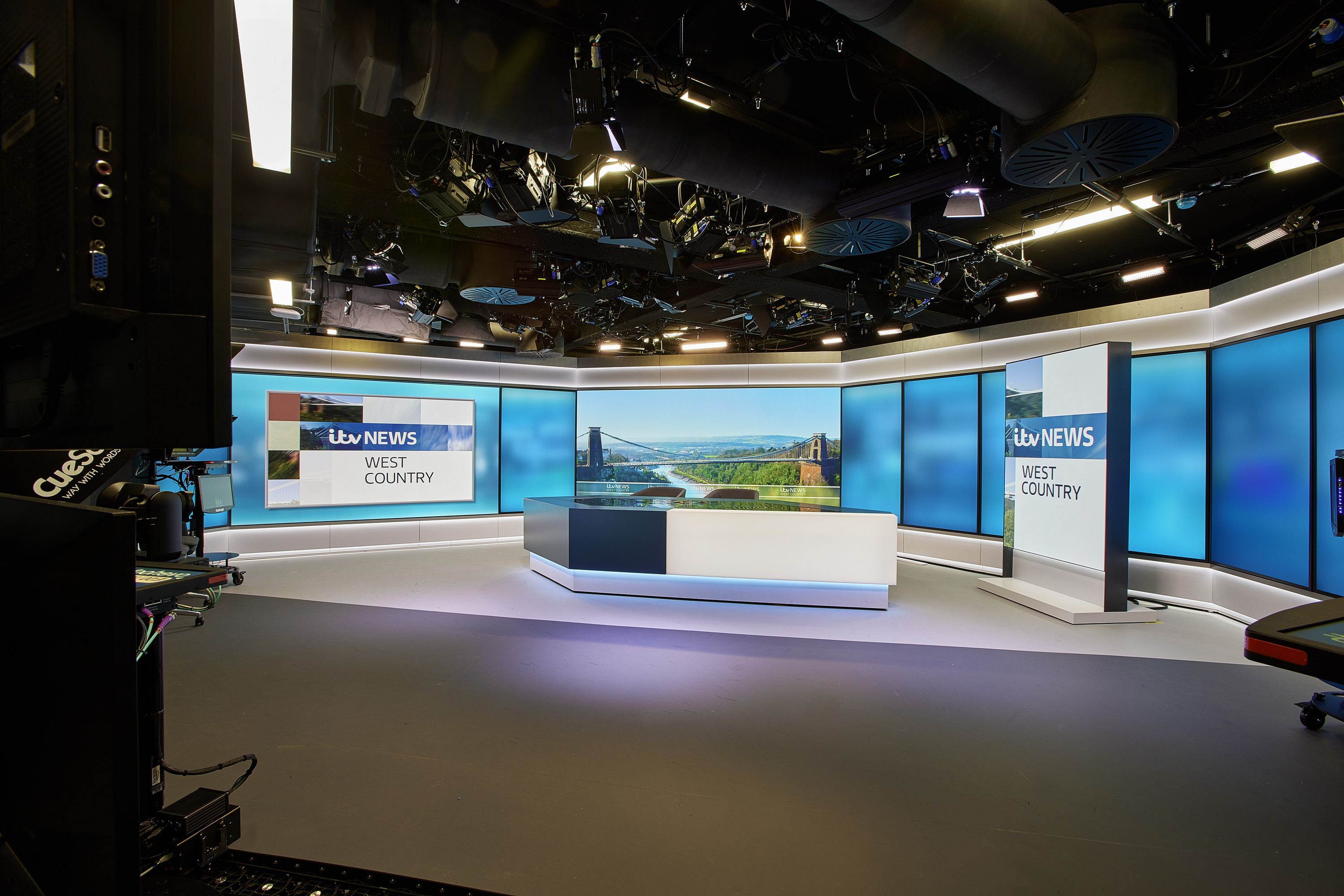 A modern television news studio set with a sleek, professional design. In the centre, a curved news desk with a white and dark grey base features an integrated screen displaying a landscape image. Behind the desk, a wide wall of large blue-toned display panels wraps around the studio, creating a bright, high-tech backdrop.  On the right side, a vertical freestanding screen shows the text “itv NEWS WEST COUNTRY” in bold lettering. A larger horizontal screen on the left wall repeats the same branding. The studio floor is smooth and grey, with subtle lighting creating soft reflections.  Above, the ceiling is open, revealing an array of lighting rigs, cameras, cables and ventilation systems, all arranged in a dense technical grid. Long linear light fixtures run parallel across the ceiling, illuminating the set evenly. On the far left edge of the image, part of a large black camera or broadcast equipment rig is visible, reinforcing the production environment. The overall space feels polished, controlled and designed for live broadcasting.