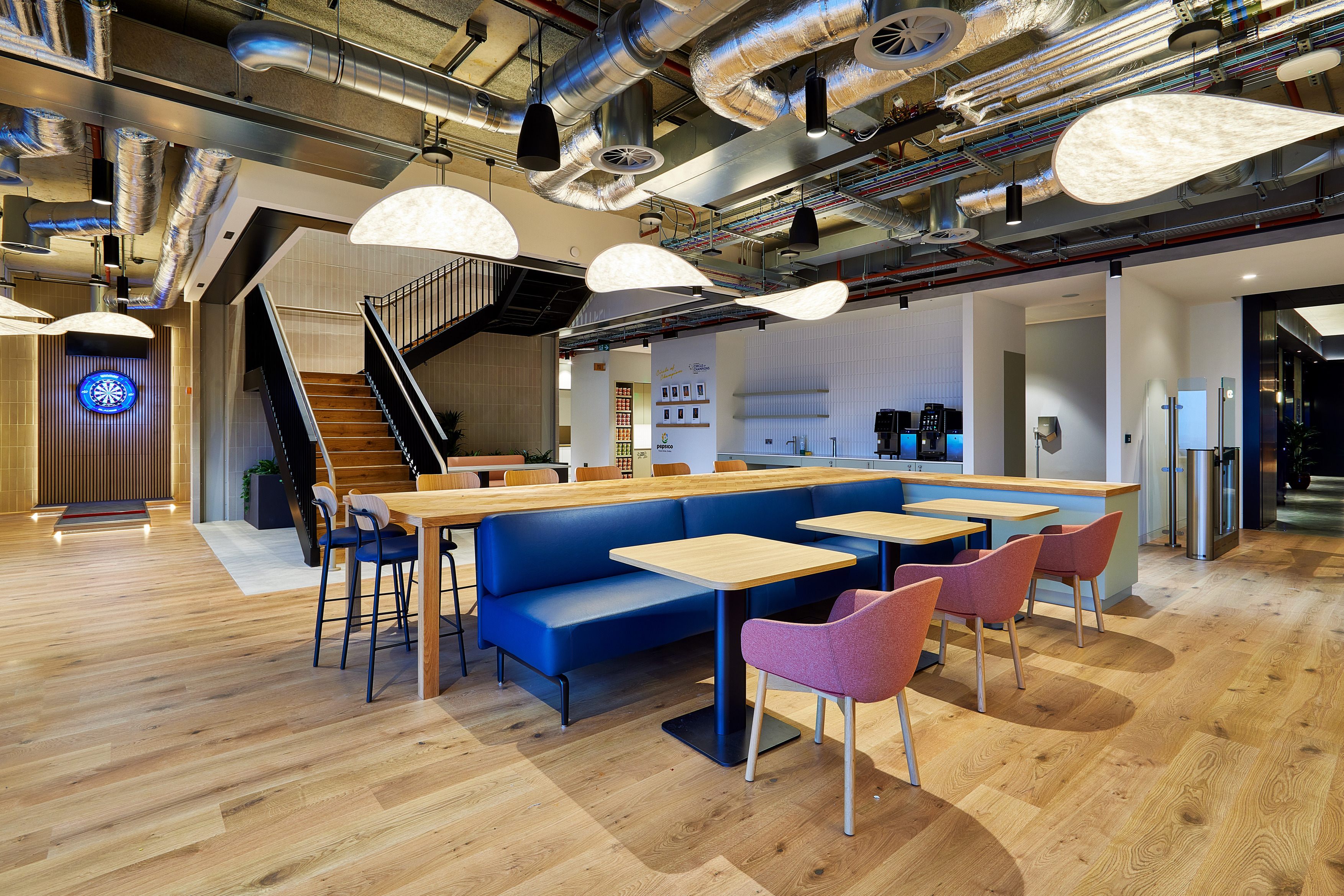 Open plan office breakout area with long communal tables, blue upholstered bench seating and pink chairs on a light wood floor. Exposed ductwork and cables run across the ceiling with large pendant lights. A staircase with black railings leads to an upper level, while a kitchenette with coffee machines sits to the rear.