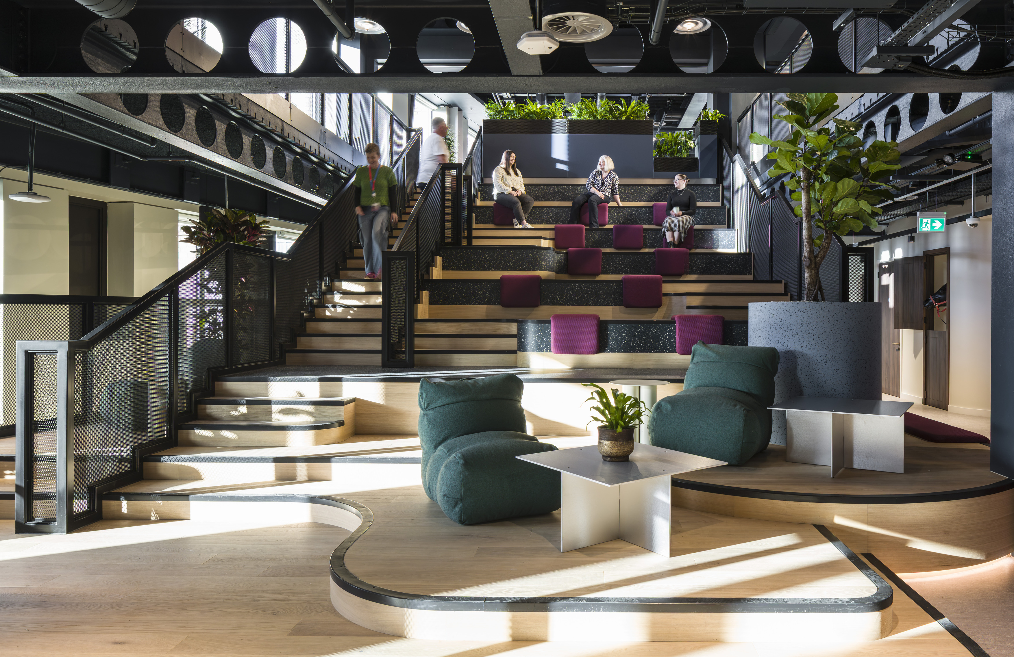 A multi level workplace space featuring tiered seating with purple cushions, surrounded by planting and framed by black steel beams. Curved wooden platforms with low lounge chairs and small white tables create informal seating on the lower level. Several people sit and walk through the space. Large planters and full height windows bring in greenery and natural light.