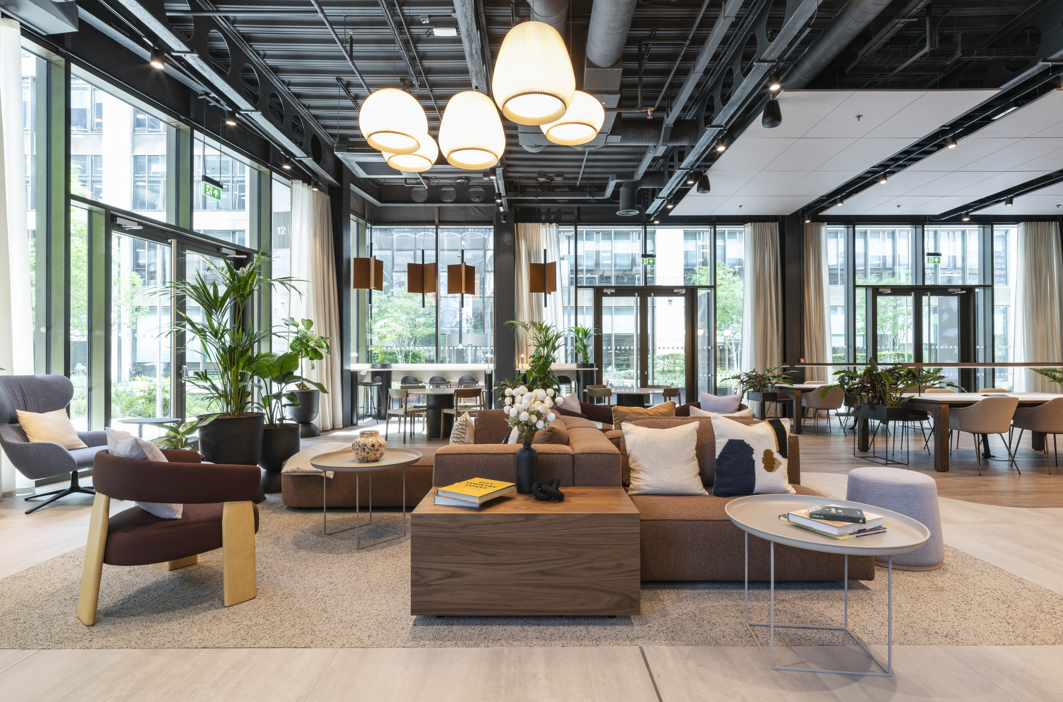 Modern office lounge area featuring a mix of contemporary furniture, including modular sofas, armchairs, and round coffee tables arranged on a textured rug. The space has large floor-to-ceiling windows for natural light, complemented by pendant lighting and exposed ceiling elements for an industrial aesthetic. Indoor plants add greenery, while neutral tones with wood accents create a warm, inviting atmosphere suitable for collaborative work and informal meetings