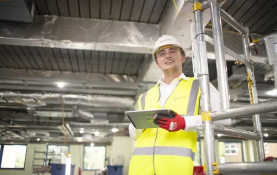Person in a high visibility yellow vest and white hard hat stands beside metal scaffolding in a construction site, holding a tablet with red work gloves, with exposed ductwork and ceiling services visible overhead.