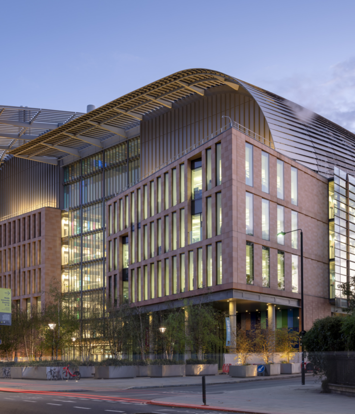 Architecturally distinctive building with a curved roof and large glass windows spanning multiple storeys. The exterior combines glass and brick materials, creating a visually striking contrast. In the foreground, trees and streetlights line the street, adding context to the urban setting