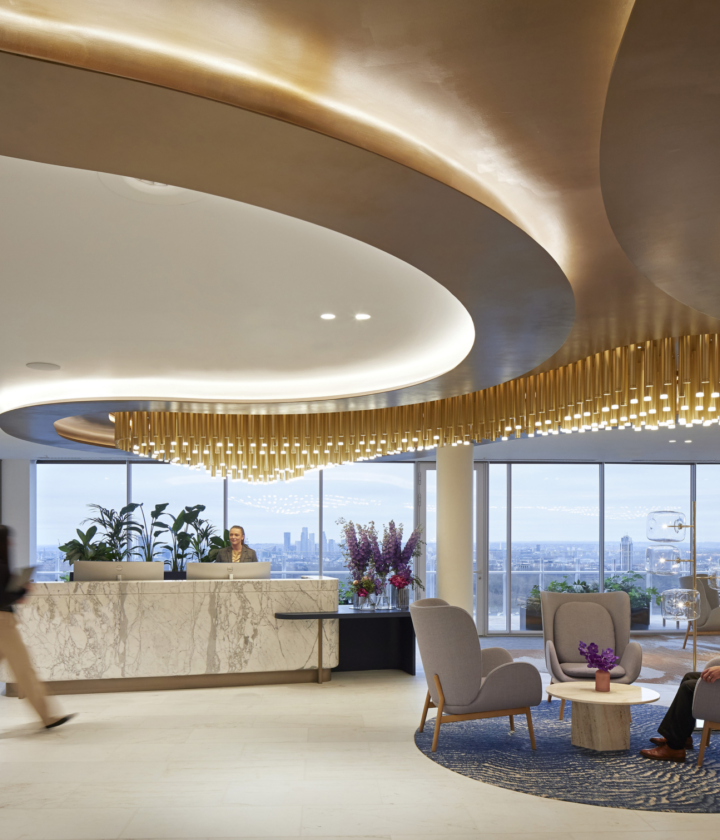 Modern office lobby with a refined and elegant design, featuring a reception desk on the left and a large curved ceiling installation adorned with hanging golden elements. The space includes a seating area with two chairs and a small table topped with flowers, creating a welcoming atmosphere. Large windows at the back offer views of an urban skyline, while the overall design blends luxury with contemporary minimalism.