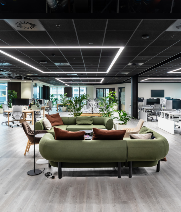 An open plan office space. Green sofas are prominent in the centre, with desks along the right and meeting pods on the left.