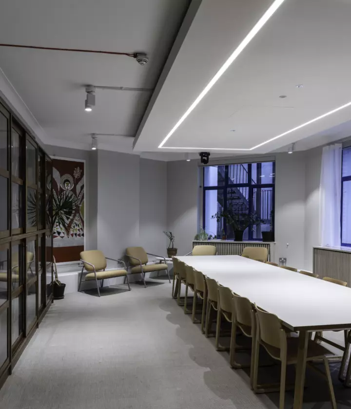 Conference room with a long white table, wooden chairs, bookshelves, and large windows with sheer curtains.