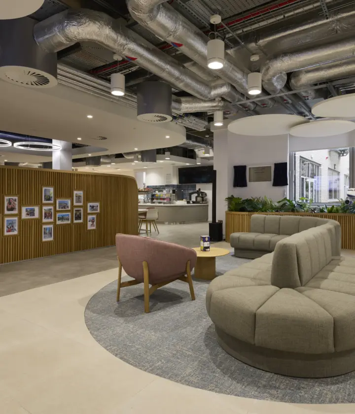 Modern office breakout space with curved upholstered seating arranged around a circular rug, a lounge chair and coffee table in the foreground. A timber feature wall displaying framed photographs separates the area from a café and kitchen space in the background. Exposed services and acoustic ceiling panels create an industrial feel, while planting and large windows bring natural light into the interior.