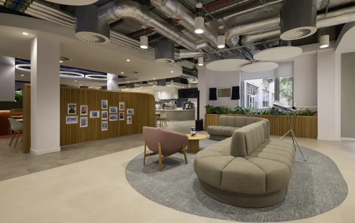 Modern office breakout space with curved upholstered seating arranged around a circular rug, a lounge chair and coffee table in the foreground. A timber feature wall displaying framed photographs separates the area from a café and kitchen space in the background. Exposed services and acoustic ceiling panels create an industrial feel, while planting and large windows bring natural light into the interior.