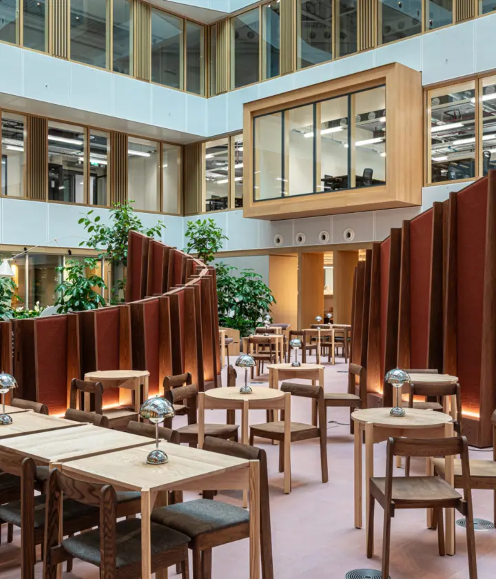 Modern open-plan office atrium with high ceilings and large windows. The space features wooden tables and chairs arranged in clusters separated by curved freestanding partitions in a reddish-brown fabric. Several green plants add a natural element throughout the area. A large digital screen is visible on the right wall and an enclosed glass meeting room is suspended above the ground level. The overall design combines wood, glass and greenery for a contemporary and inviting atmosphere.