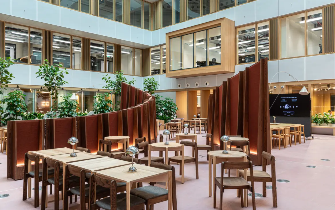 Modern open-plan office atrium with high ceilings and large windows. The space features wooden tables and chairs arranged in clusters separated by curved freestanding partitions in a reddish-brown fabric. Several green plants add a natural element throughout the area. A large digital screen is visible on the right wall and an enclosed glass meeting room is suspended above the ground level. The overall design combines wood, glass and greenery for a contemporary and inviting atmosphere.