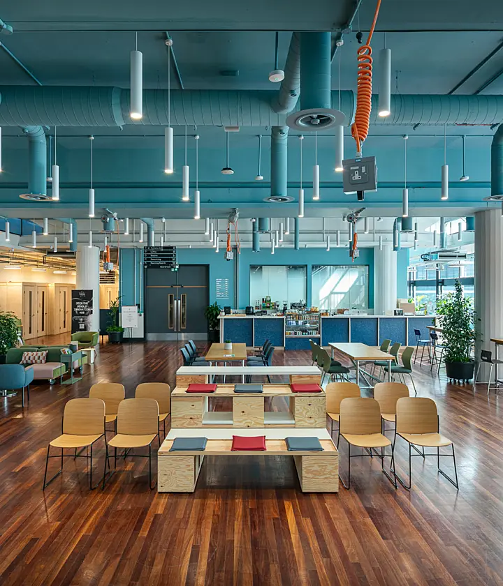 Open-plan workplace lounge and collaboration space with polished wood flooring, tiered timber seating, tables and chairs arranged for informal meetings, and a café counter at the rear. The interior features a teal-painted exposed ceiling with visible ductwork, suspended lighting and hanging power outlets. Large windows along one side bring in natural light, while plants are placed throughout the space.
