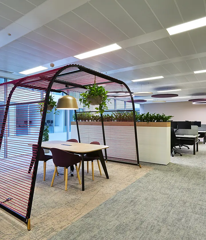 Modern open-plan office space featuring a semi-enclosed meeting pod with red slatted screening around a table and four chairs. The office includes desk workstations, glass-fronted meeting rooms, large planters with greenery and soft overhead lighting, creating a bright and contemporary workplace environment.