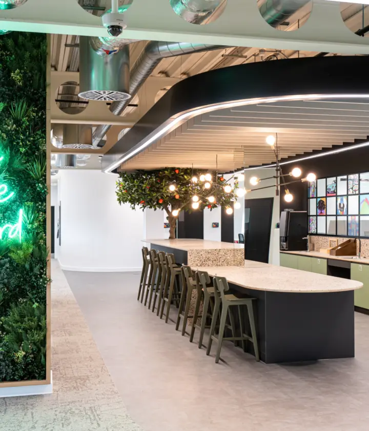 A stylish modern office kitchen with a long island counter, bar stools and overhead lighting. A vertical garden wall on the left displays neon text reading "Welcome to the Orchard." Wall-mounted cabinets and clean finishes complete the inviting, contemporary workspace.