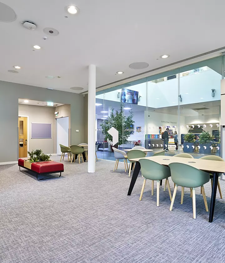 Modern open-plan office space featuring a mix of collaborative and breakout areas. The design includes a large glass partition wall creating transparency and natural light flow complemented by neutral grey carpeting and white ceilings with integrated circular acoustic panels. A long table with light green chairs provides a shared workspace while a red accent wall adds colour contrast near wooden doors. Soft seating in red and green tones is arranged around low planters for informal meetings. The fit out incorporates minimalist furniture, clean lines and biophilic elements with indoor plants creating a contemporary and welcoming environment.
