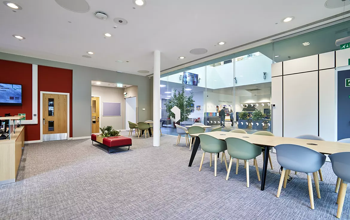 Modern open-plan office space featuring a mix of collaborative and breakout areas. The design includes a large glass partition wall creating transparency and natural light flow complemented by neutral grey carpeting and white ceilings with integrated circular acoustic panels. A long table with light green chairs provides a shared workspace while a red accent wall adds colour contrast near wooden doors. Soft seating in red and green tones is arranged around low planters for informal meetings. The fit out incorporates minimalist furniture, clean lines and biophilic elements with indoor plants creating a contemporary and welcoming environment.