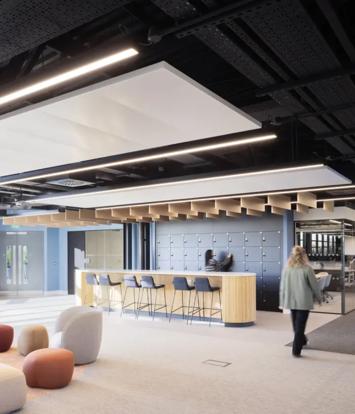 A bright open office space with soft seating in various colours arranged near full height windows. A bar height counter with stools sits in the centre of the room with a wall of lockers behind it. One person stands at the counter and another walks across the space. The ceiling features exposed black panels and linear lighting. A glass meeting room is visible on the right.