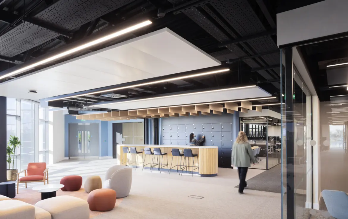 A bright open office space with soft seating in various colours arranged near full height windows. A bar height counter with stools sits in the centre of the room with a wall of lockers behind it. One person stands at the counter and another walks across the space. The ceiling features exposed black panels and linear lighting. A glass meeting room is visible on the right.
