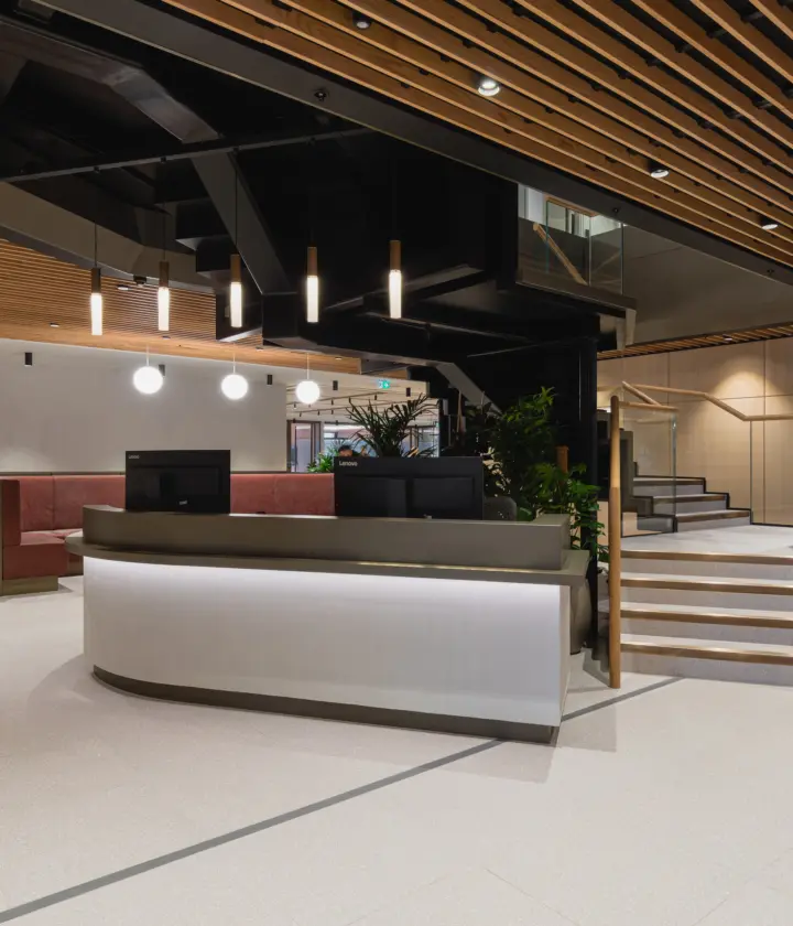 Modern office reception area with a curved white reception desk, light stone flooring and a striking timber slatted ceiling. Black structural features and pendant lights create contrast, while staircases with glass balustrades lead to upper levels. Built-in seating and indoor planting add warmth to the contemporary lobby space.