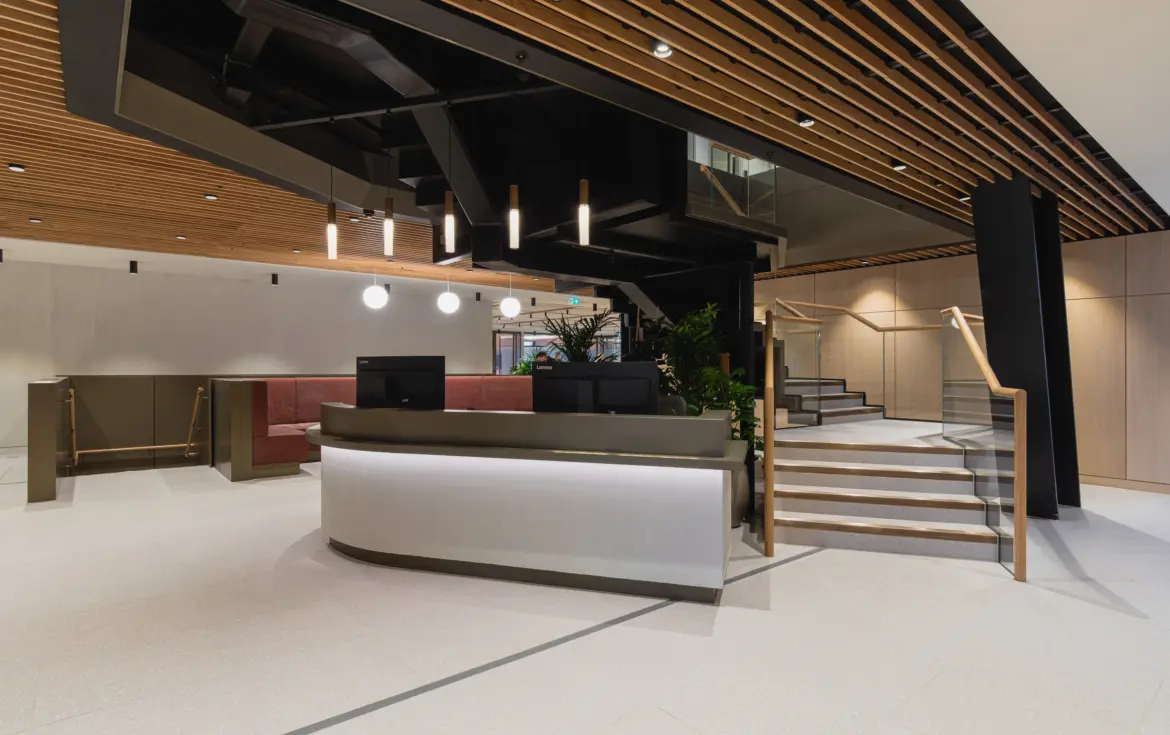 Modern office reception area with a curved white reception desk, light stone flooring and a striking timber slatted ceiling. Black structural features and pendant lights create contrast, while staircases with glass balustrades lead to upper levels. Built-in seating and indoor planting add warmth to the contemporary lobby space.