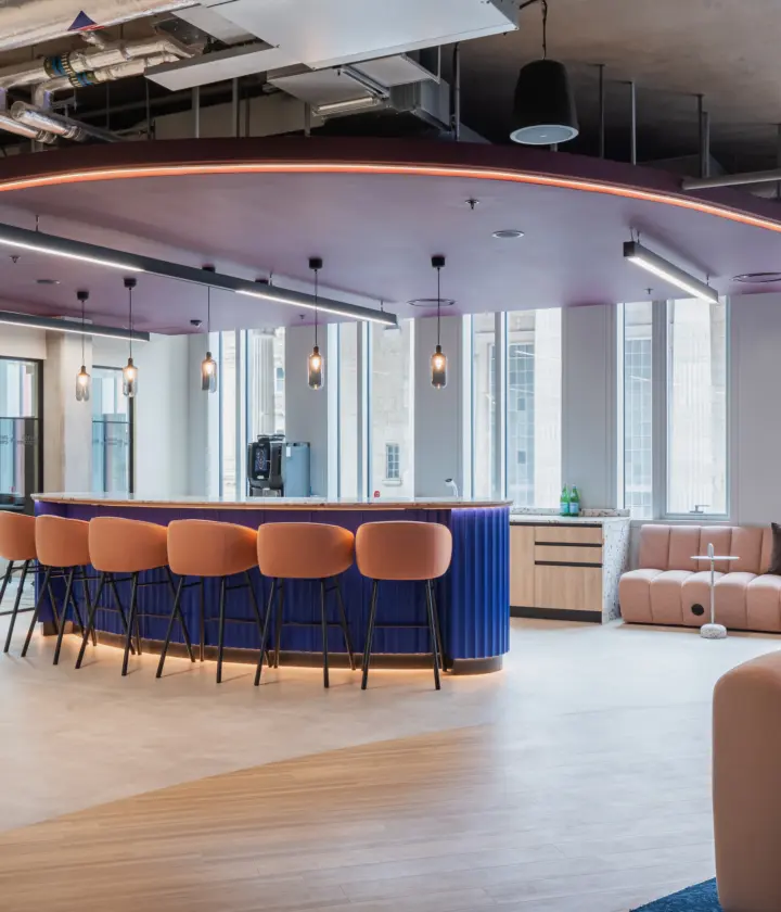 Modern office breakout space with a curved blue bar and peach-upholstered stools beneath a suspended ceiling feature with integrated lighting. Large windows bring in natural light, while soft seating areas with curved sofas and small tables create informal meeting and collaboration spaces. Exposed services and contemporary finishes give the interior a stylish, open-plan feel.