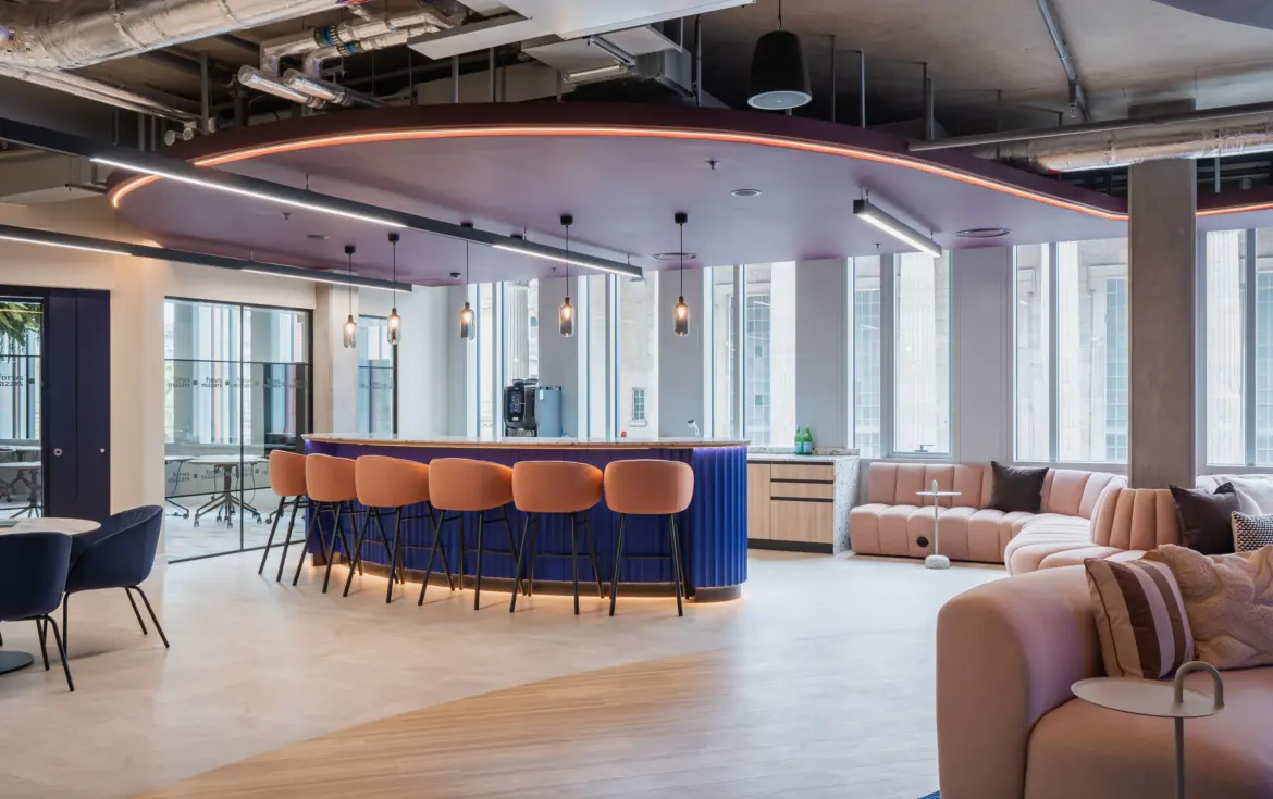 Modern office breakout space with a curved blue bar and peach-upholstered stools beneath a suspended ceiling feature with integrated lighting. Large windows bring in natural light, while soft seating areas with curved sofas and small tables create informal meeting and collaboration spaces. Exposed services and contemporary finishes give the interior a stylish, open-plan feel.