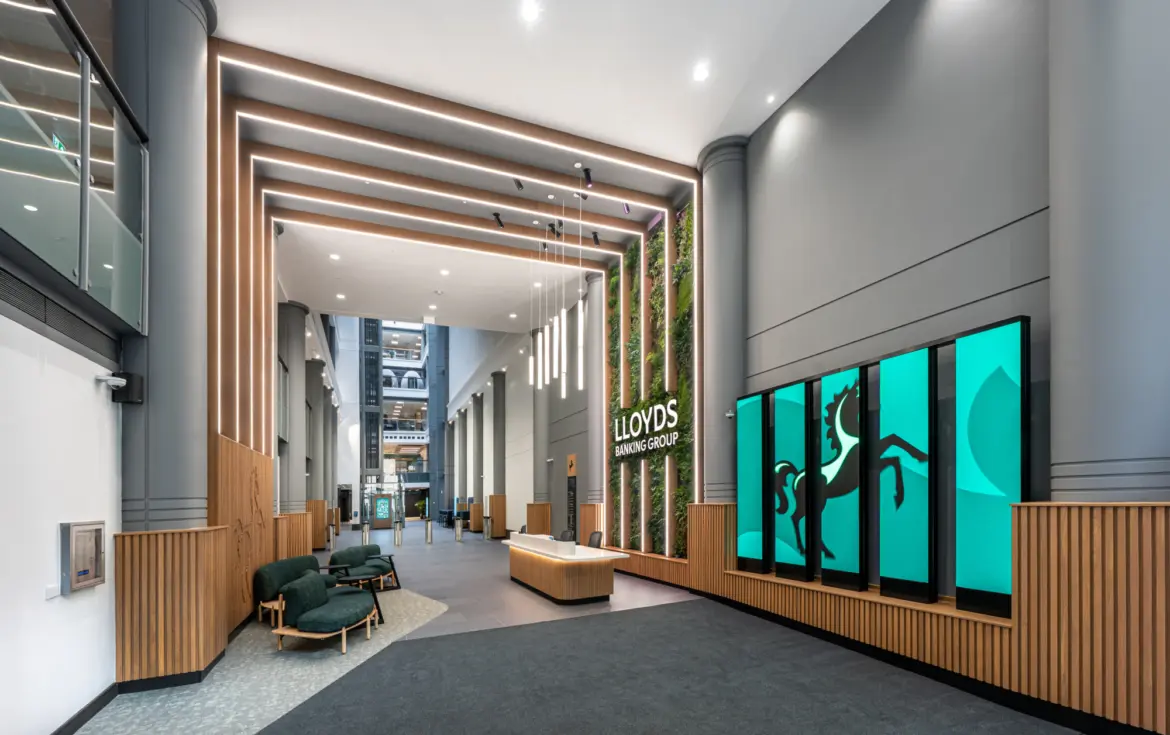 A spacious modern lobby with high ceilings and tall grey walls featuring vertical wooden slats. A series of suspended linear lights hangs above a central reception desk made of light wood. To the right, a green living wall surrounds the Lloyds Banking Group logo. Next to it, large digital screens display the company’s black horse symbol on a teal background. Soft seating with dark green upholstery sits to the left, and the far end of the space opens into a bright atrium with additional architectural details.