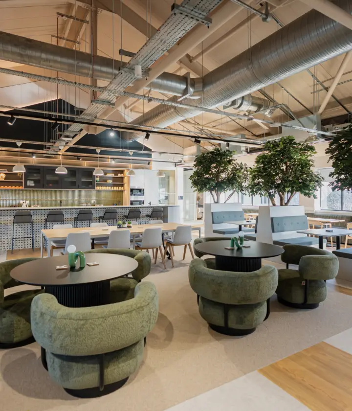 Modern open-plan office breakout and café area featuring a central kitchen island with bar seating, round tables surrounded by green upholstered lounge chairs and large indoor trees in white planters. Exposed ceiling services, light wood flooring and floor-to-ceiling windows create a bright, contemporary workspace.