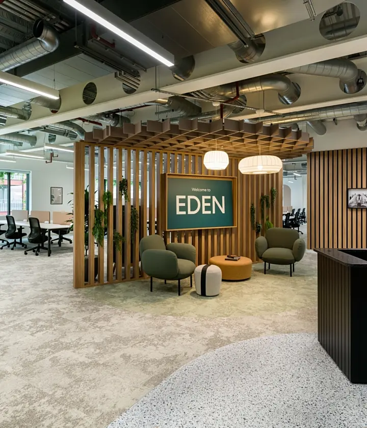 Bright, modern office reception area with an open-plan layout. In the centre, a wooden slatted feature structure frames a green sign reading “Welcome to EDEN”, with soft seating including armchairs and a round ottoman arranged beneath warm pendant lights. To the right, a dark reception desk sits against a wall of vertical wooden slats displaying framed black-and-white photos. To the left, a workspace with desks and office chairs is visible through large windows. The ceiling features exposed ductwork, pipes and linear lighting, while the floor combines carpet and polished concrete finishes, giving the space a contemporary, industrial feel.