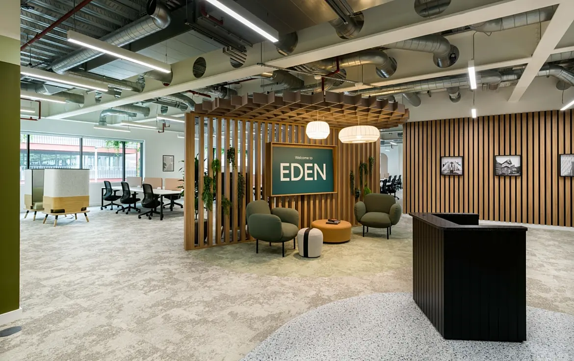 Bright, modern office reception area with an open-plan layout. In the centre, a wooden slatted feature structure frames a green sign reading “Welcome to EDEN”, with soft seating including armchairs and a round ottoman arranged beneath warm pendant lights. To the right, a dark reception desk sits against a wall of vertical wooden slats displaying framed black-and-white photos. To the left, a workspace with desks and office chairs is visible through large windows. The ceiling features exposed ductwork, pipes and linear lighting, while the floor combines carpet and polished concrete finishes, giving the space a contemporary, industrial feel.
