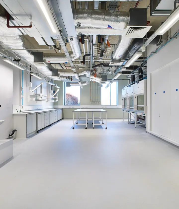 Bright, empty laboratory interior with white floors and cabinets, a central worktable and exposed ceiling services including ducts, pipes and lighting, facing a window at the far end.