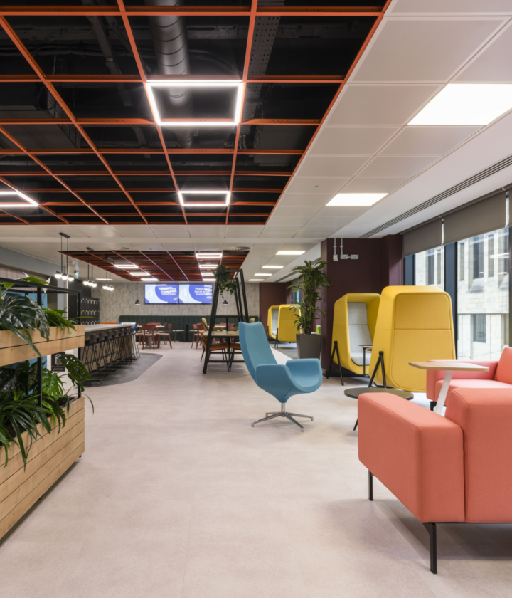 The image shows a modern office fit out and office design with a variety of seating options and decor. On the left are plants in wooden planters, a blue chair sits in the centre and a pink couch is on the right. In the background are yellow and green enclosed seating areas, tables, chairs and large windows that let in natural light. The ceiling features an orange grid pattern with recessed lighting.