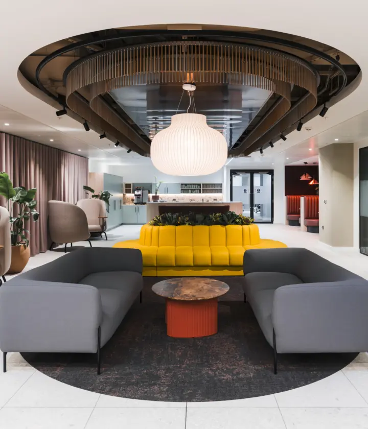 Contemporary office lounge and reception area featuring curved grey sofas arranged around a circular coffee table, with a bright yellow banquette and integrated planting at the centre. A large decorative pendant light hangs beneath a sculptural ceiling feature with timber detailing and black accents. Soft lounge chairs line the perimeter beside full-height curtains, while glazed meeting rooms and a refreshment area are visible in the background. The space combines light stone flooring, warm neutral tones and modern furnishings to create a welcoming hospitality-inspired workplace environment.