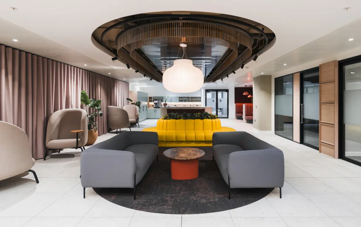 Contemporary office lounge and reception area featuring curved grey sofas arranged around a circular coffee table, with a bright yellow banquette and integrated planting at the centre. A large decorative pendant light hangs beneath a sculptural ceiling feature with timber detailing and black accents. Soft lounge chairs line the perimeter beside full-height curtains, while glazed meeting rooms and a refreshment area are visible in the background. The space combines light stone flooring, warm neutral tones and modern furnishings to create a welcoming hospitality-inspired workplace environment.