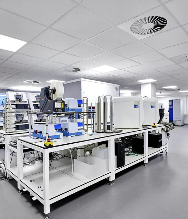 Modern laboratory space featuring automated testing and analytical equipment arranged on white workbenches within a large open-plan room. The laboratory has grey flooring, white ceilings with recessed lighting and ventilation units, large windows along one side and clear walkways between workstations. Clean, bright and organised, the space is designed for scientific research and technical analysis.