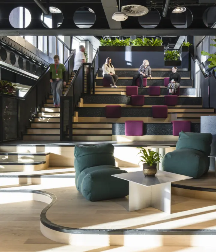 A multi level workplace space featuring tiered seating with purple cushions, surrounded by planting and framed by black steel beams. Curved wooden platforms with low lounge chairs and small white tables create informal seating on the lower level. Several people sit and walk through the space. Large planters and full height windows bring in greenery and natural light.