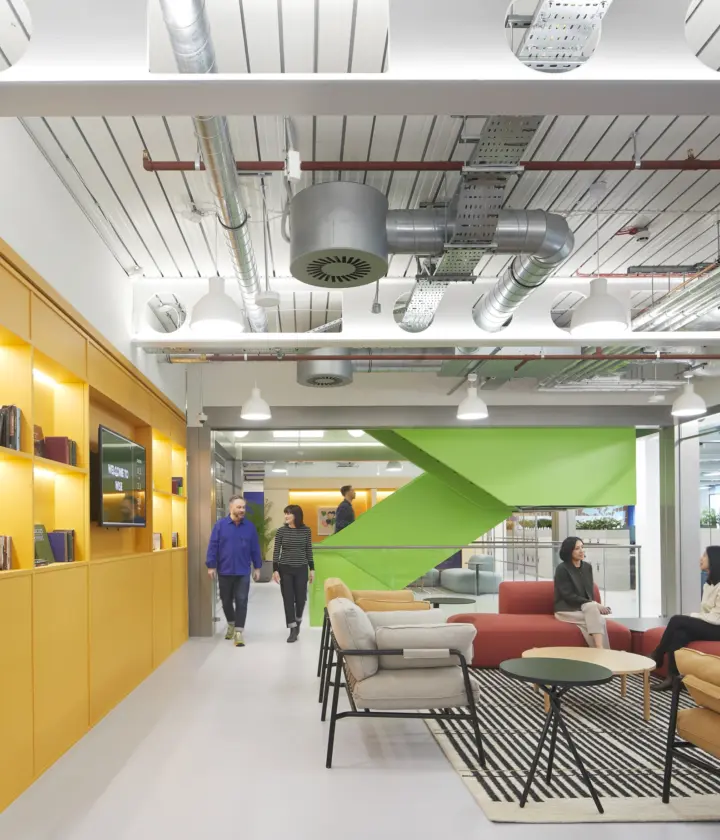 Open plan office fit out with a modern breakout area featuring colourful sofas and armchairs, built-in shelving, exposed ceiling services and suspended lighting, with staff using the space for informal meetings within a contemporary office design.