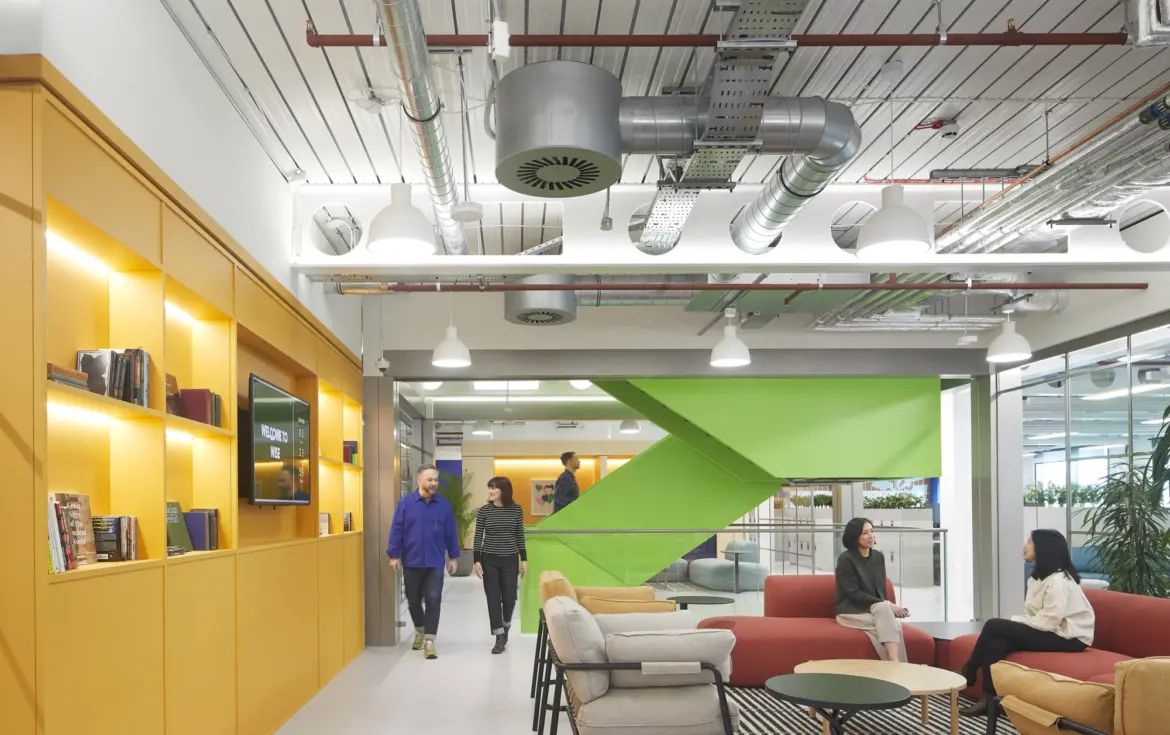 Open plan office fit out with a modern breakout area featuring colourful sofas and armchairs, built-in shelving, exposed ceiling services and suspended lighting, with staff using the space for informal meetings within a contemporary office design.