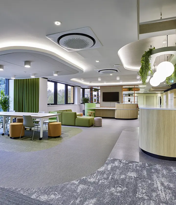 Spacious modern office breakout area with a curved white ceiling, circular air vents and feature lighting. A central wooden counter sits beside a kitchenette with green cabinetry and integrated appliances. To the left, a collaborative workspace includes a white table with chairs and soft seating in green and mustard tones, set on a carpeted zone. Large windows along the wall bring in natural light and overlook greenery outside. Decorative elements include vertical green wall panels, indoor plants and a suspended ceiling feature with hanging foliage, creating a biophilic design throughout.