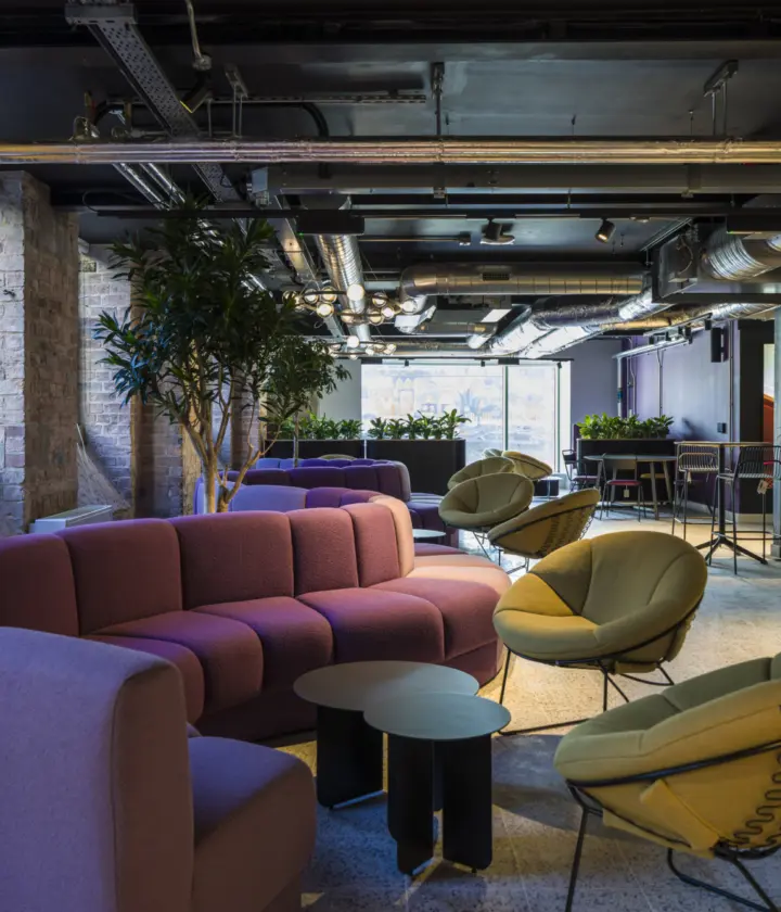 Modern office lounge with an industrial aesthetic featuring exposed brick walls and visible ductwork on a black ceiling. The space includes curved modular sofas in muted purple tones, paired with round black side tables. Two green lounge chairs with wire bases add contrast. The flooring is light terrazzo, and large potted plants provide greenery throughout. In the background, there are café-style tables and chairs, complemented by a vibrant abstract wall mural. Natural light filters through large windows, enhancing the open and collaborative atmosphere.