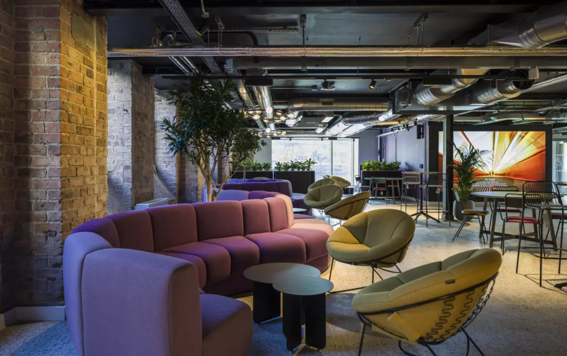 Modern office lounge with an industrial aesthetic featuring exposed brick walls and visible ductwork on a black ceiling. The space includes curved modular sofas in muted purple tones, paired with round black side tables. Two green lounge chairs with wire bases add contrast. The flooring is light terrazzo, and large potted plants provide greenery throughout. In the background, there are café-style tables and chairs, complemented by a vibrant abstract wall mural. Natural light filters through large windows, enhancing the open and collaborative atmosphere.