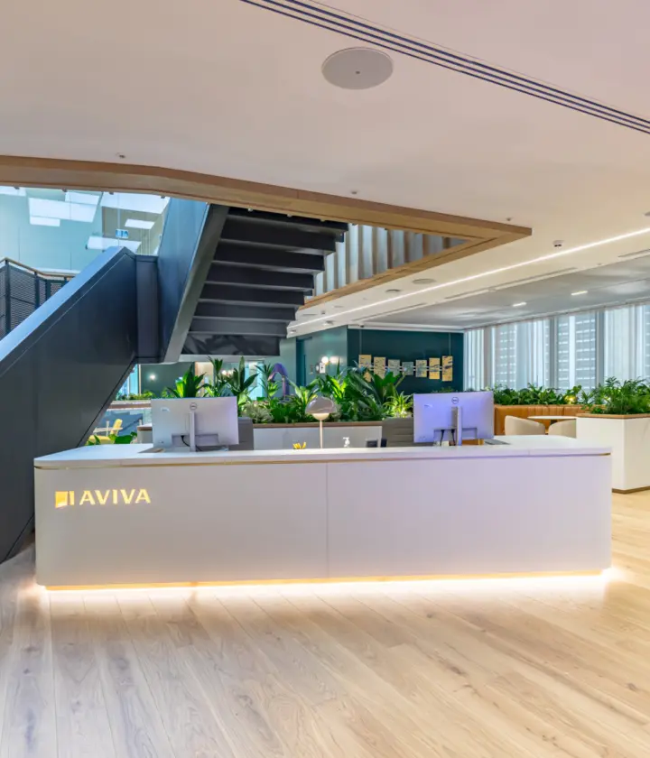 A bright modern office reception area with a white desk featuring the Aviva logo, two computer monitors and soft underlighting. Behind the desk are large planters filled with greenery. A wide wooden staircase with a black metal balustrade rises to an upper floor. The space has light wooden flooring, floor to ceiling windows with blinds and a mix of open workspaces and plants.