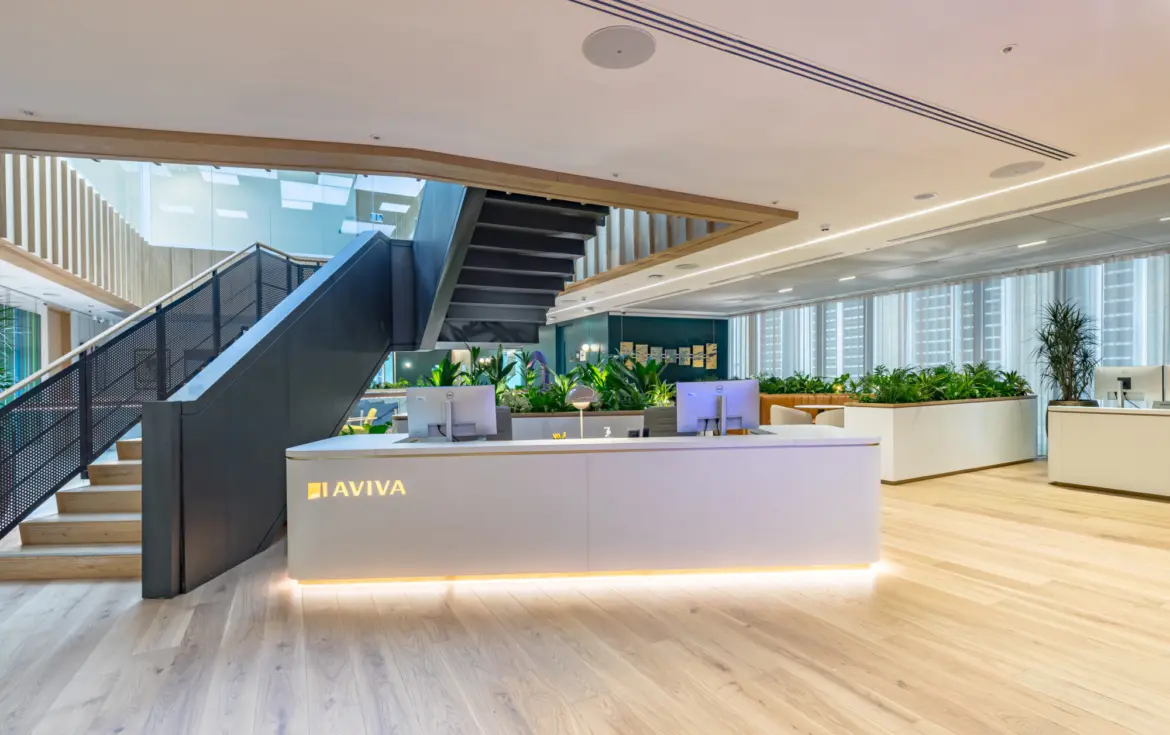 A bright modern office reception area with a white desk featuring the Aviva logo, two computer monitors and soft underlighting. Behind the desk are large planters filled with greenery. A wide wooden staircase with a black metal balustrade rises to an upper floor. The space has light wooden flooring, floor to ceiling windows with blinds and a mix of open workspaces and plants.