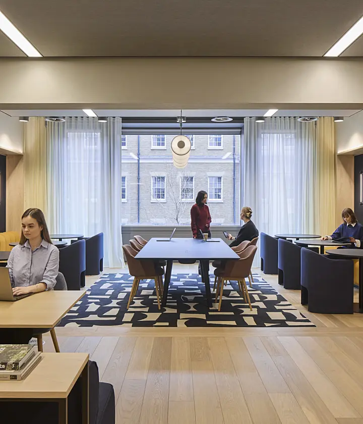 ￼￼Modern office lounge and reception area with light wood flooring, dark ceiling panels, exposed services and linear lighting. A curved blue sofa, armchairs and coffee tables sit on a patterned rug in the foreground, while several people work and meet throughout the open-plan space. Large windows, indoor planting and neutral finishes create a bright, contemporary interior.
