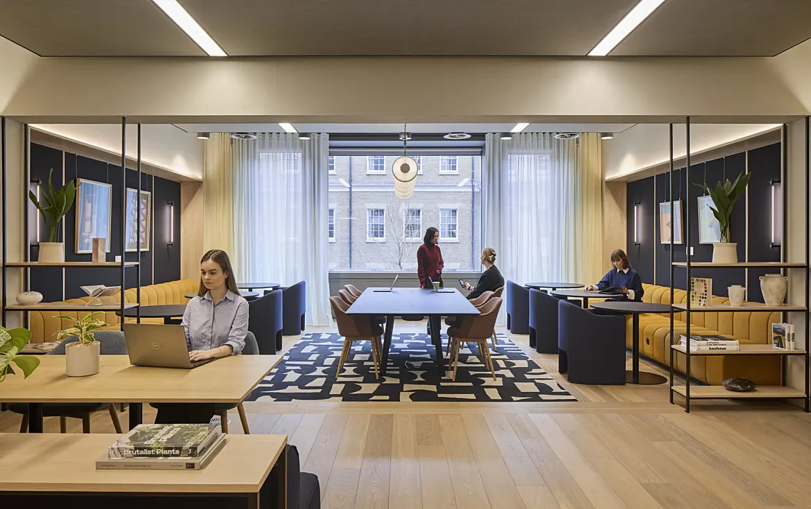 ￼￼Modern office lounge and reception area with light wood flooring, dark ceiling panels, exposed services and linear lighting. A curved blue sofa, armchairs and coffee tables sit on a patterned rug in the foreground, while several people work and meet throughout the open-plan space. Large windows, indoor planting and neutral finishes create a bright, contemporary interior.