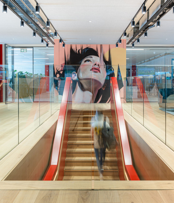 The image shows a modern office fit out and office design. It features an open-plan layout with wooden flooring and glass railings. A staircase with red handrails leads to a lower level, bordered by glass panels. At the top of the stairs is a large mural of a woman's face, adding an artistic element to the space. The ceiling has track lighting and there are several seating areas with contemporary furniture in the background. The overall design highlights openness, light and modern aesthetics.
