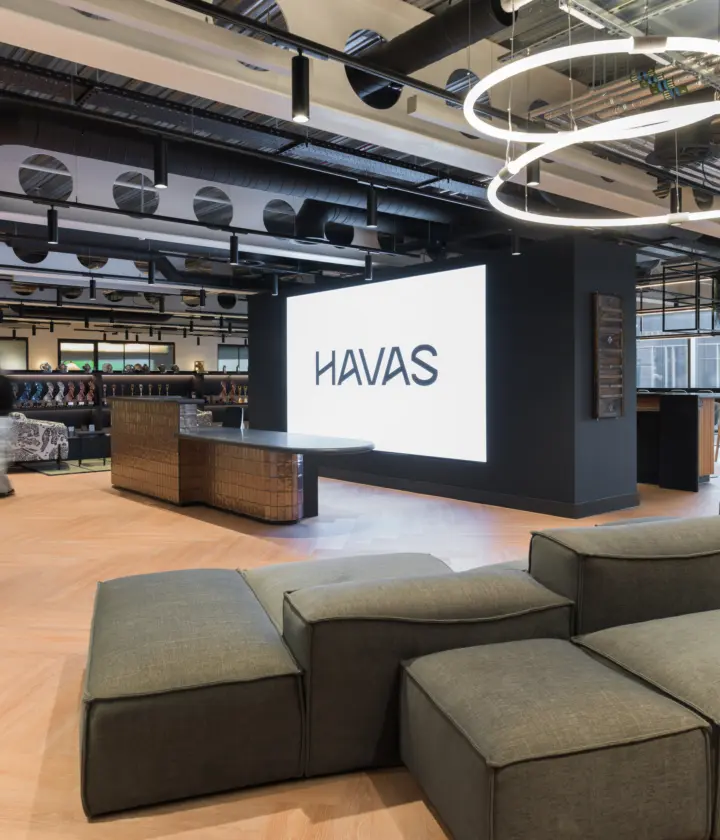 Modern open-plan office lounge with light wood flooring, exposed ceiling services and circular LED light fixtures. A large illuminated HAVAS logo is displayed on a central feature wall beside a curved reception desk. Modular grey seating fills the foreground, while booths, tables and workspaces extend across the bright interior. Several people are visible using the space near floor-to-ceiling windows.