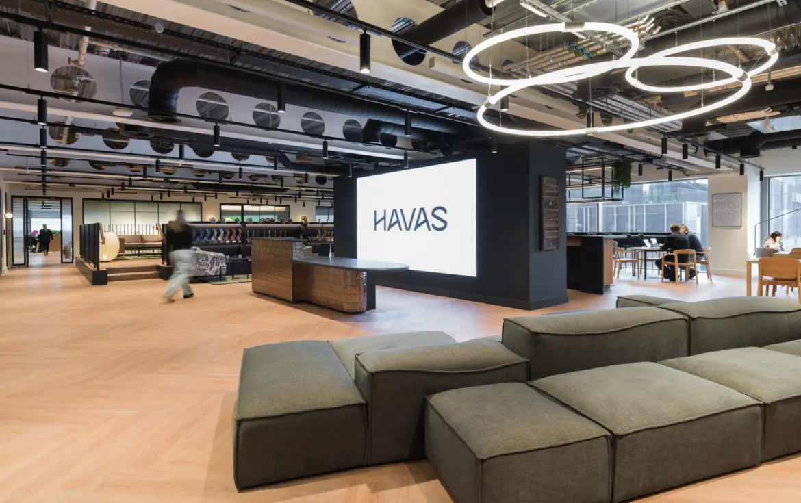 Modern open-plan office lounge with light wood flooring, exposed ceiling services and circular LED light fixtures. A large illuminated HAVAS logo is displayed on a central feature wall beside a curved reception desk. Modular grey seating fills the foreground, while booths, tables and workspaces extend across the bright interior. Several people are visible using the space near floor-to-ceiling windows.