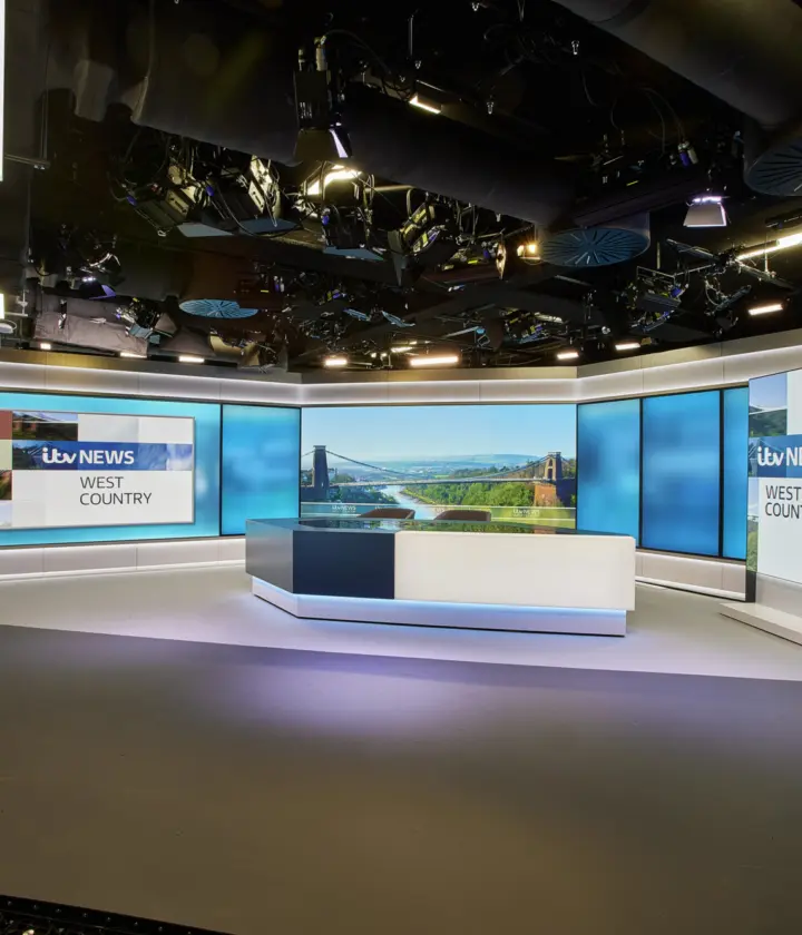 A modern television news studio set with a sleek, professional design. In the centre, a curved news desk with a white and dark grey base features an integrated screen displaying a landscape image. Behind the desk, a wide wall of large blue-toned display panels wraps around the studio, creating a bright, high-tech backdrop.  On the right side, a vertical freestanding screen shows the text “itv NEWS WEST COUNTRY” in bold lettering. A larger horizontal screen on the left wall repeats the same branding. The studio floor is smooth and grey, with subtle lighting creating soft reflections.  Above, the ceiling is open, revealing an array of lighting rigs, cameras, cables and ventilation systems, all arranged in a dense technical grid. Long linear light fixtures run parallel across the ceiling, illuminating the set evenly. On the far left edge of the image, part of a large black camera or broadcast equipment rig is visible, reinforcing the production environment. The overall space feels polished, controlled and designed for live broadcasting.